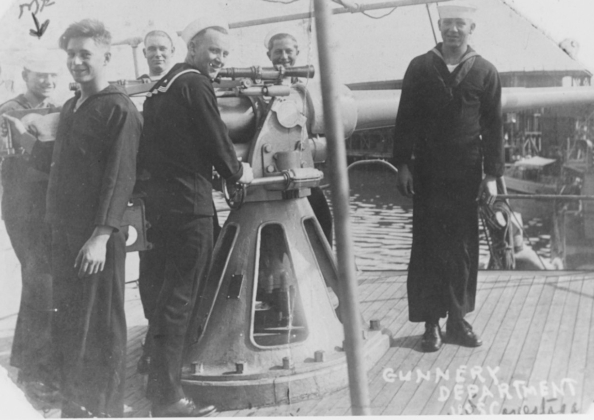 Six sailors gathered around a 3 inch 50 caliber gun on a ship's deck, smiling and looking at the camera. 